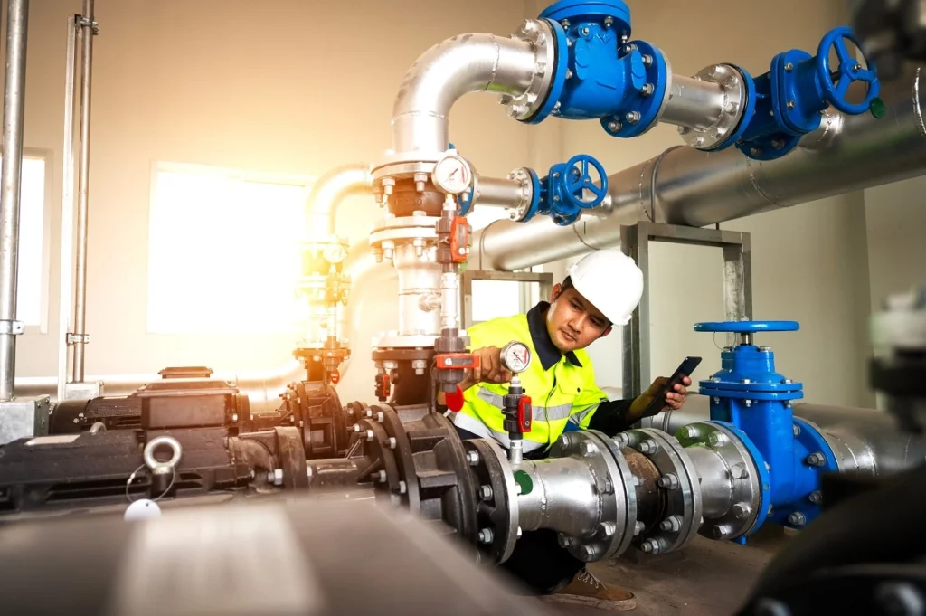 Technician in safety gear checking gauges and valves on large industrial pumps and pipelines during routine maintenance