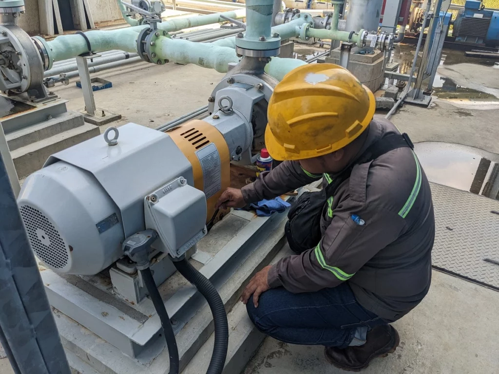 Maintenance worker wearing a yellow hard hat inspecting and servicing an industrial pump motor at an outdoor facility