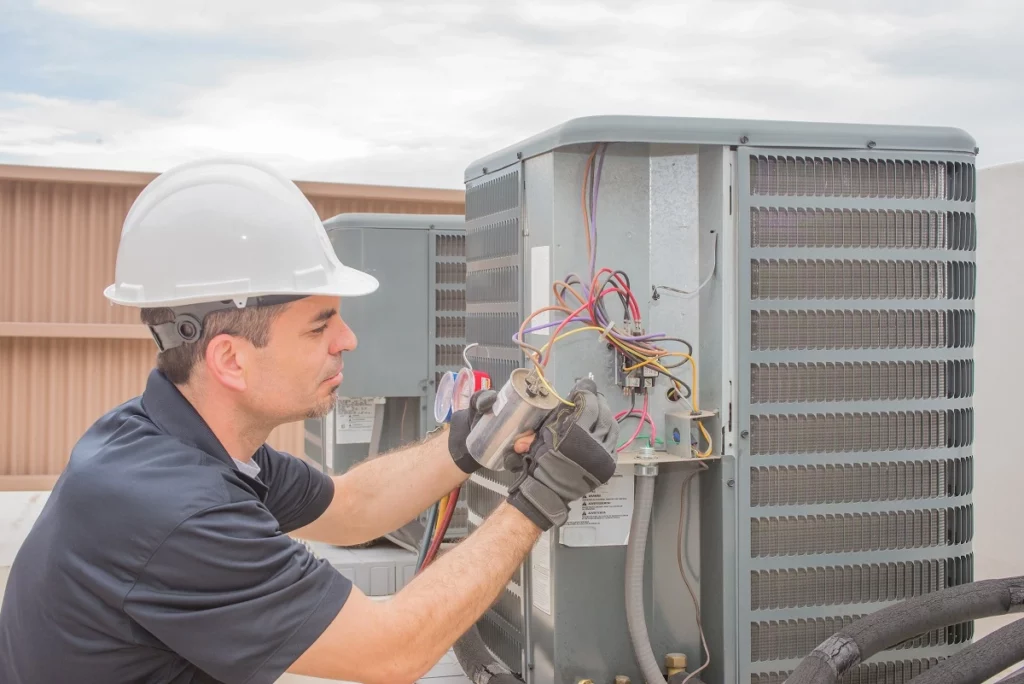 HVAC technician wearing safety gear inspects and repairs a heat pump system, checking wiring and electrical components near the unit nameplate
